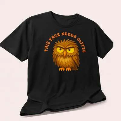 Cartoon grumpy owl with text 'This Face Needs Coffee' in brown, yellow, and orange — printed on a unisex t-shirt (angled view)