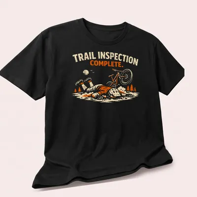 Playful illustration of a mountain biker with the text 'Trail Inspection Complete' in orange and cream tones. — printed on a unisex t-shirt (angled view)