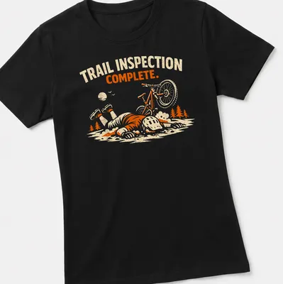 Playful illustration of a mountain biker with the text 'Trail Inspection Complete' in orange and cream tones. — printed on a women's shirt